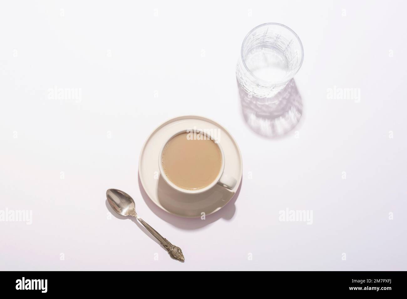 Tazza da caffè, bicchiere d'acqua e cucchiaio su tavolo bianco alla luce del sole. Vista dall'alto, disposizione piatta. Foto Stock