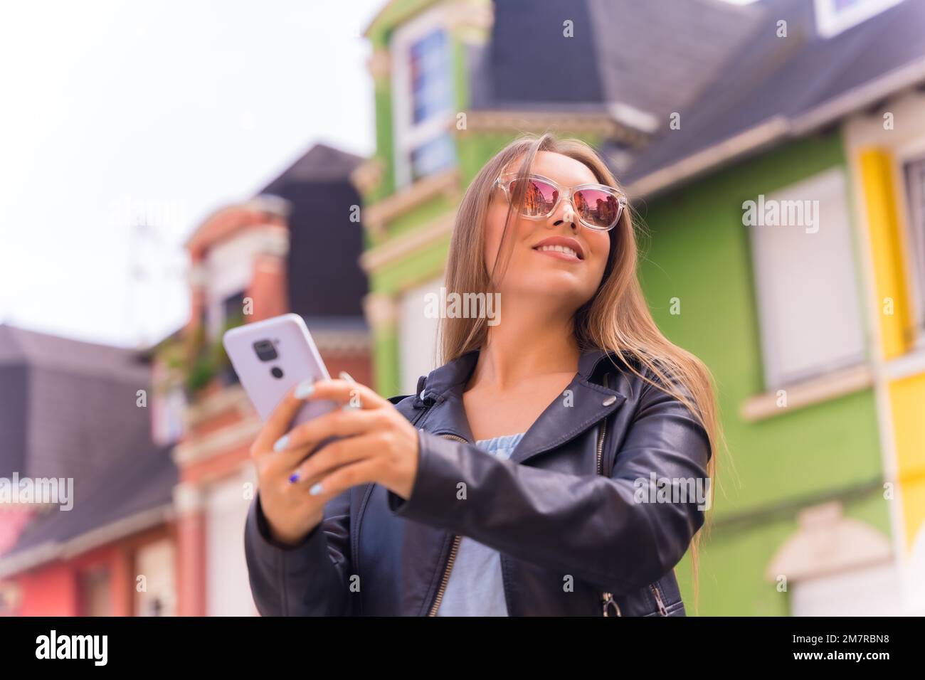 Giovane donna bionda in giacca di pelle e occhiali da sole sorridente con il mobile, dietro la facciata colorata Foto Stock