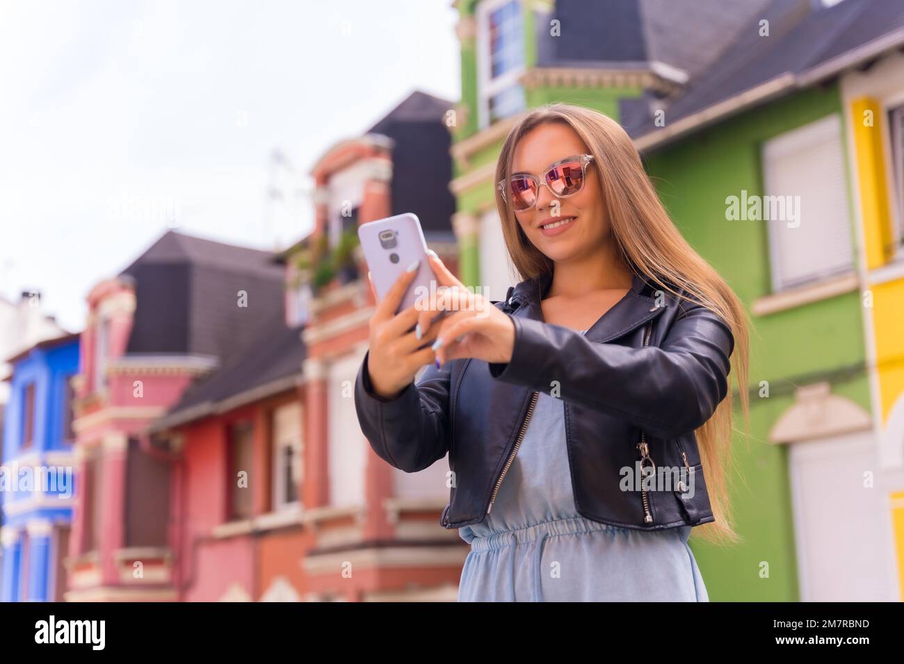 Giovane donna bionda in giacca di pelle e occhiali da sole sorridente con il mobile, dietro la facciata colorata Foto Stock