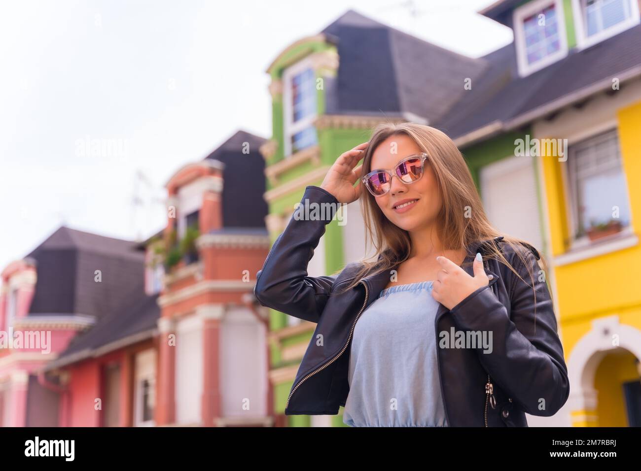 Posto di giovane donna bionda in giacca di pelle e occhiali da sole sorridenti, dietro una facciata colorata Foto Stock