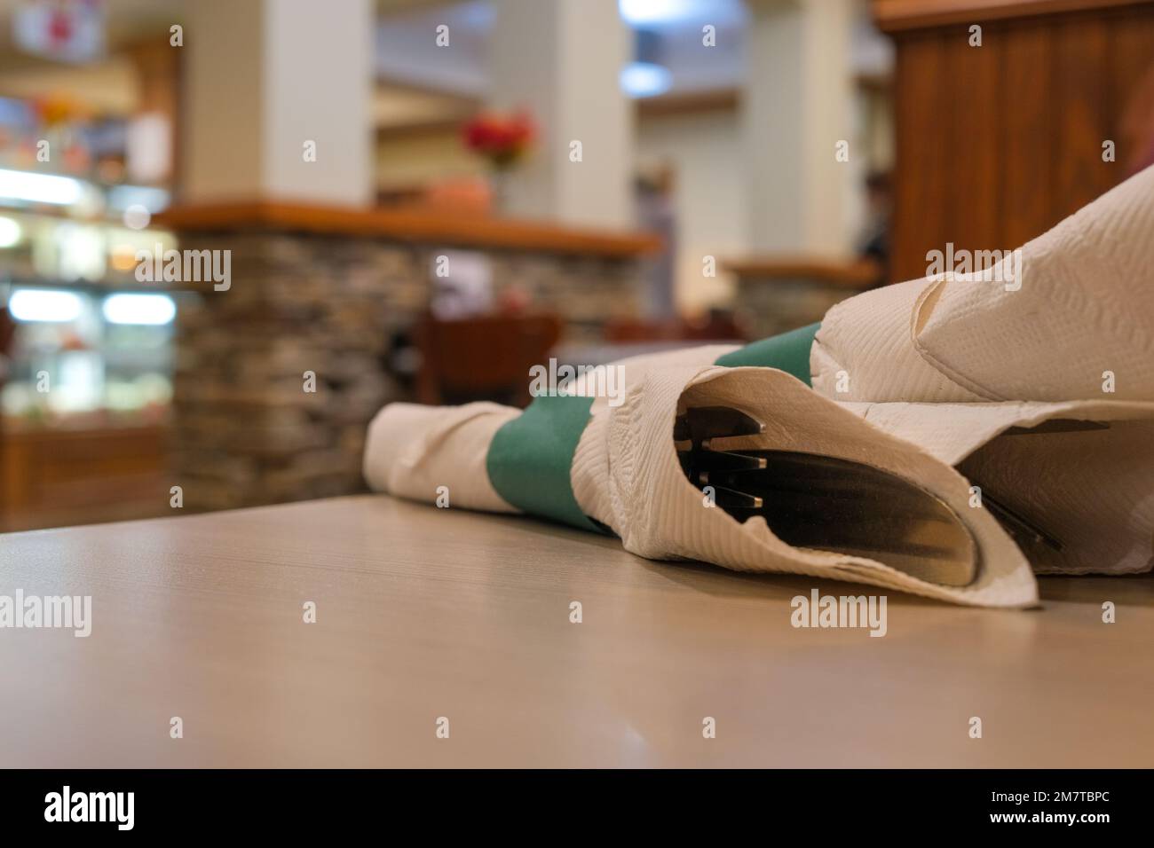 All'interno di un ristorante, le posate si trovano avvolte in un tovagliolo di carta su un tavolo. Gli utensili sono visti da vicino, con la cena sfocata sullo sfondo. Foto Stock