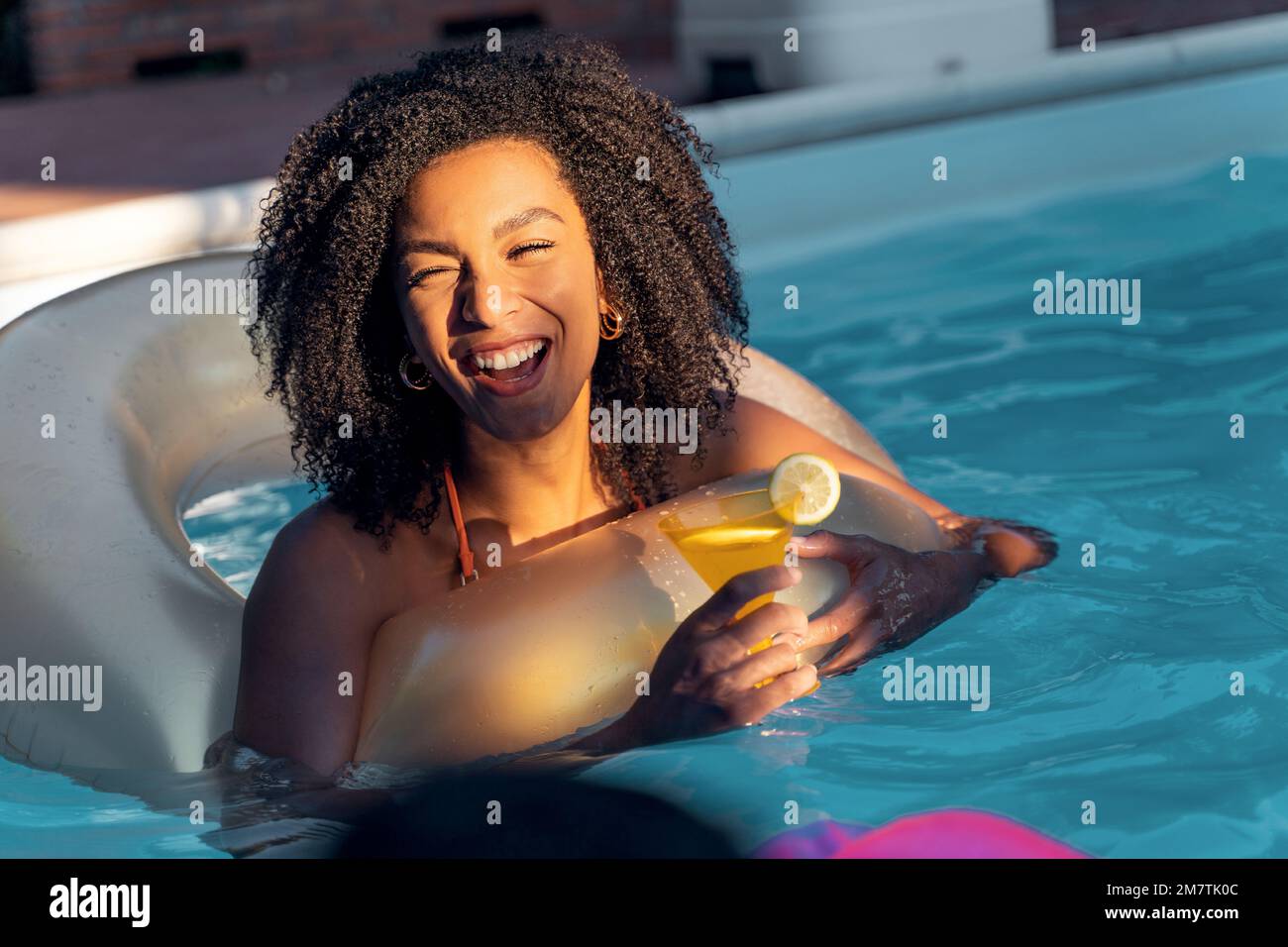 Una giovane donna multietnica con capelli ricci sta ridendo e si divertendo con le sue amiche, mentre tiene un cocktail martini che galleggia su una ciambella Foto Stock