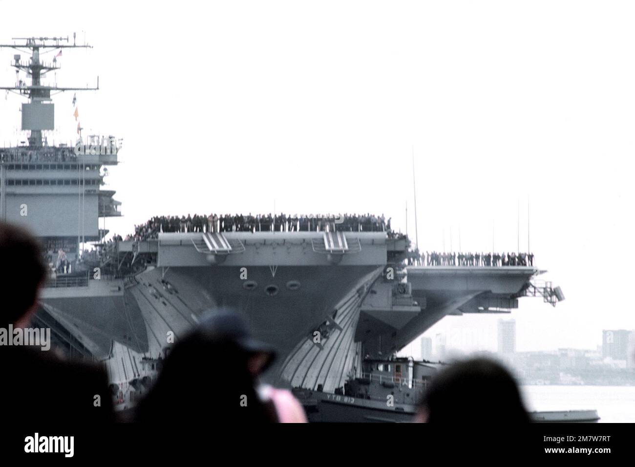Gli equipenti allineano le rotaie della portaerei a energia nucleare USS ENTERPRISE (CVN-65) mentre il grande rimorchiatore portuale USS POUGHKEEPSIE (YTB-812) spinge la portaerei in posizione di attracco. Le famiglie degli equipanti aspettano sul molo per salutare la nave. L'IMPRESA sta ritornando al porto dopo una breve crociera dopo il completamento dei recenti lavori di ristrutturazione che hanno incluso la ricostruzione della sovrastruttura. Base: Naval Air Station, Alameda Stato: California (CA) Paese: Stati Uniti d'America (USA) Foto Stock