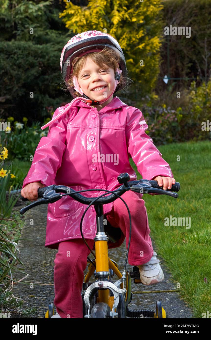 Una ragazza felice di 5 anni che cavalca un triciclo giallo giù un percorso in un cappotto impermeabile rosa in un giardino Foto Stock