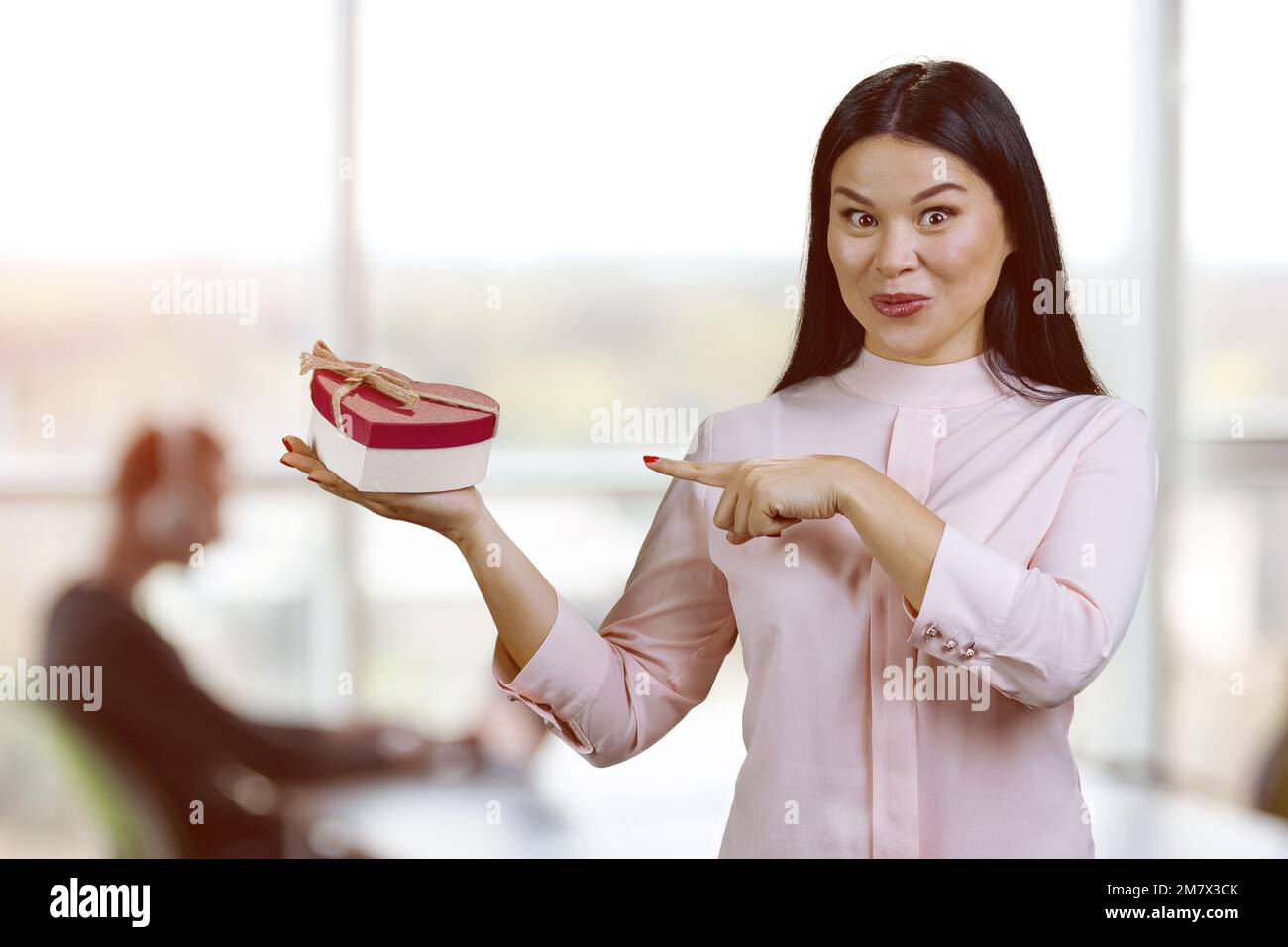 Giovane donna asiatica eccitata che mostra a forma di cuore scatola regalo. Sfondo dell'ufficio interno. Foto Stock