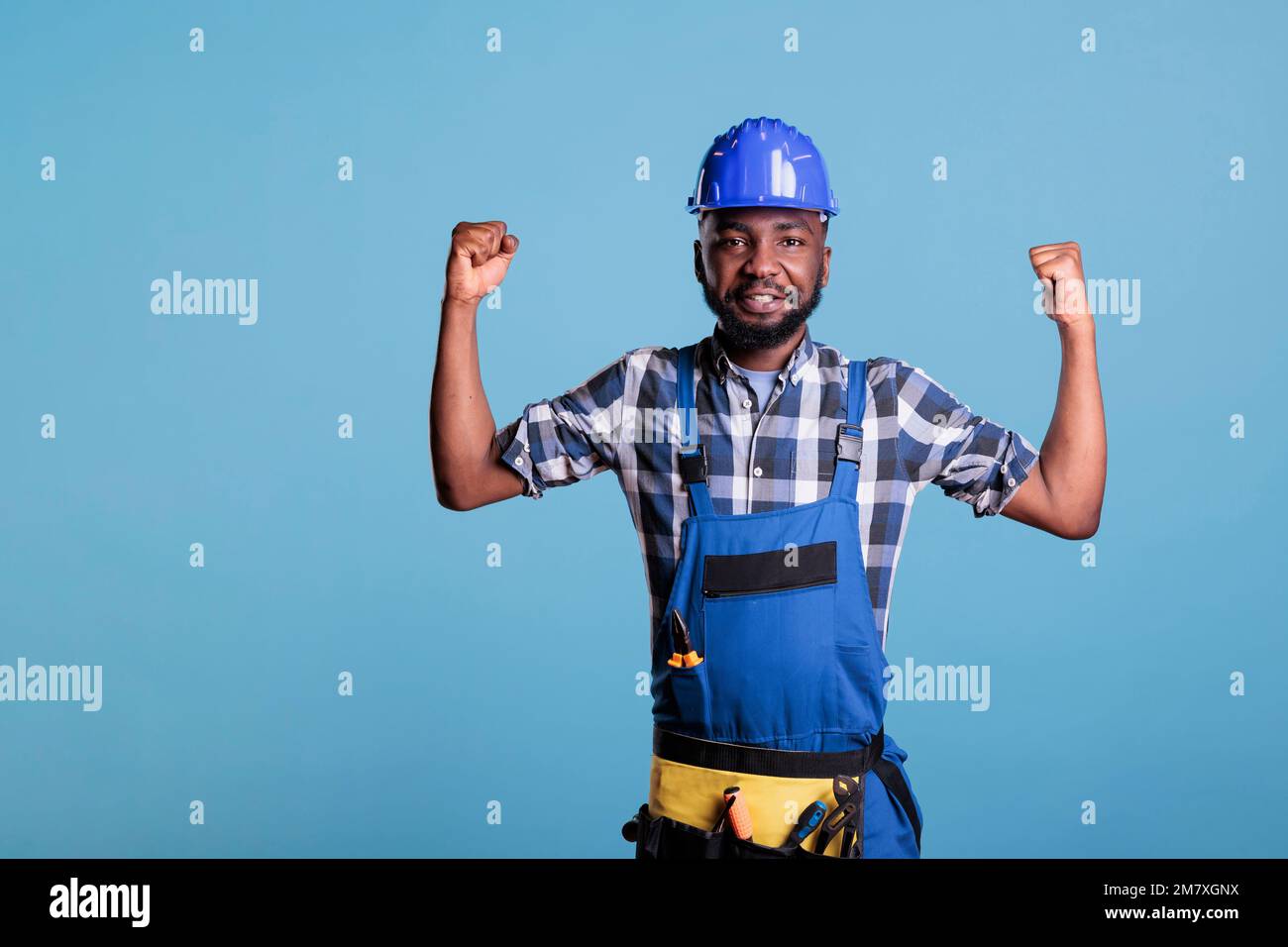 Operaio di costruzione afroamericano orgoglioso dei muscoli sviluppati del braccio dal lavoro pesante che esegue quotidiano. Il costruttore indossa tute e elmetto in studio girato su sfondo blu. Foto Stock