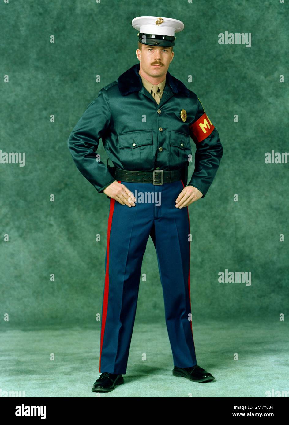 Un SGT marino. È vestito in una divisa invernale della polizia militare (MP).  Indossa pantaloni blu, una giacca marrone, un berretto bianco da caserma,  un distintivo di polizia, una fascia da braccio