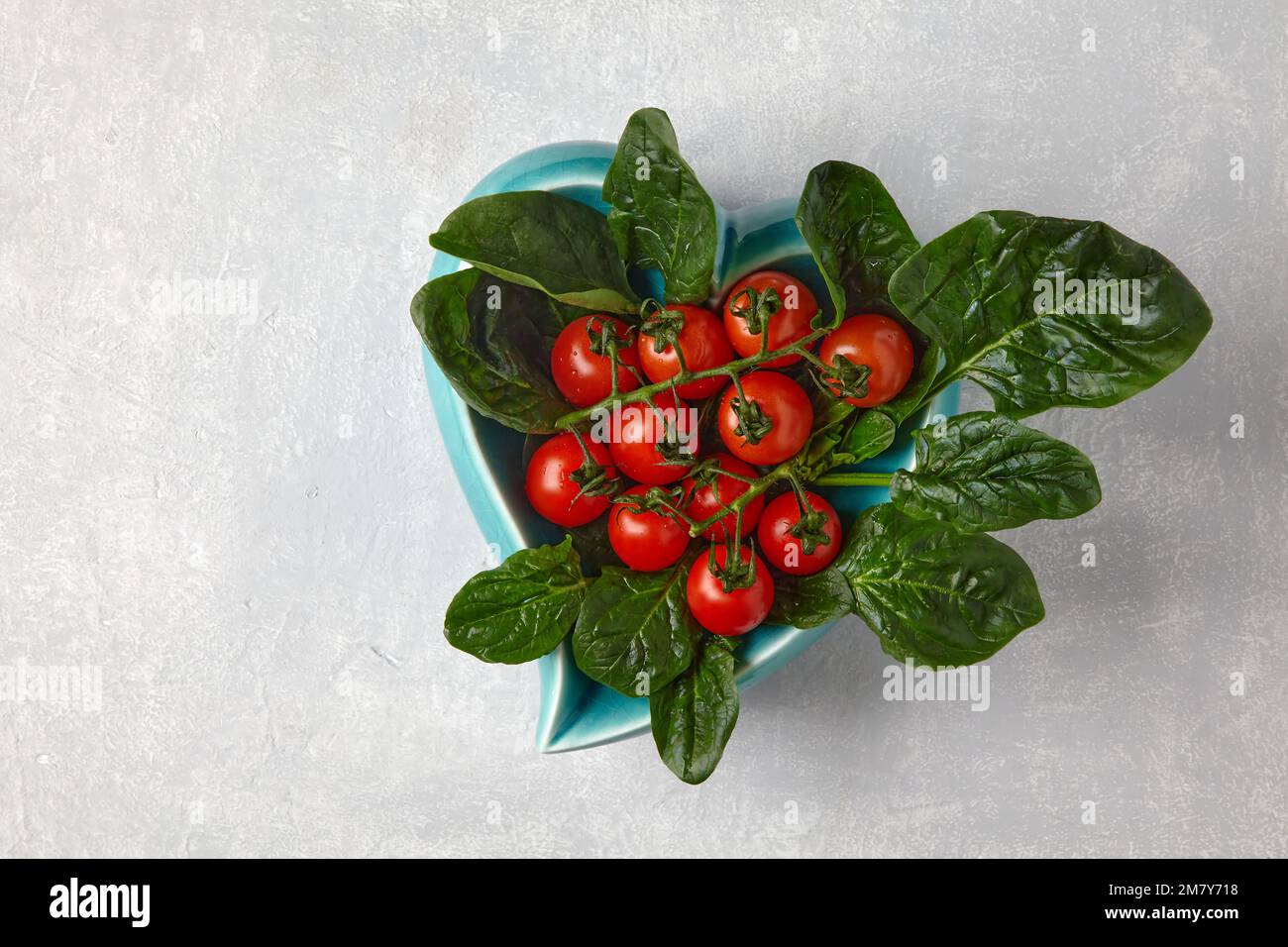 Pomodori ciliegini rossi su un ramo e foglie di spinaci verdi in una ciotola blu a forma di cuore su un tavolo di cemento chiaro Foto Stock