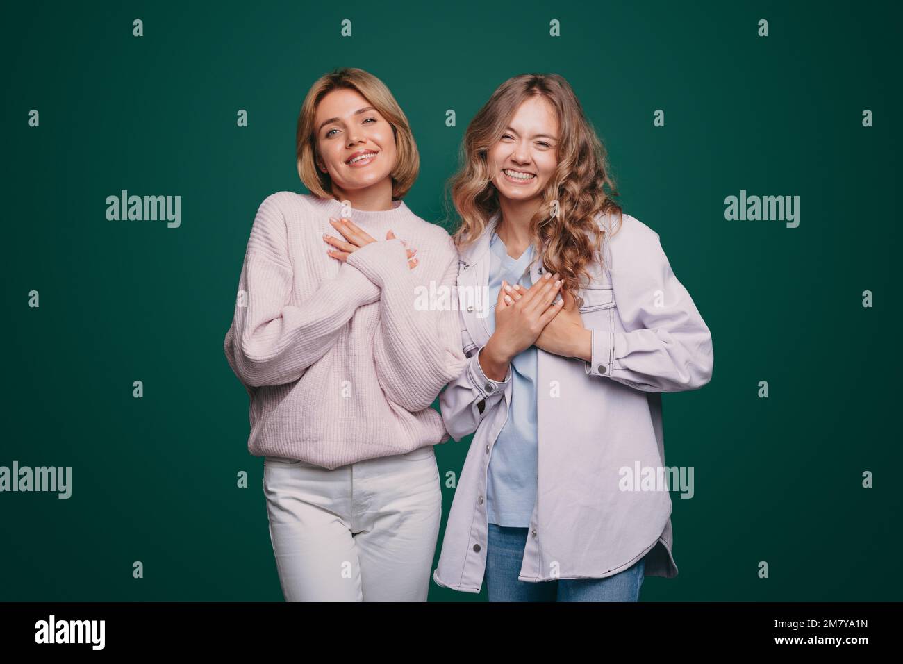 Ritratto di due giovani donne positive con emozioni sincere tenendo mani sul cuore isolato su sfondo verde Foto Stock