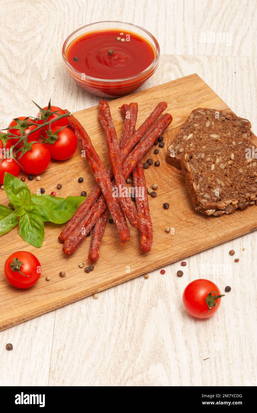 Sottili salsicce da caccia su un asse di legno decorato con pomodori e basilico Foto Stock