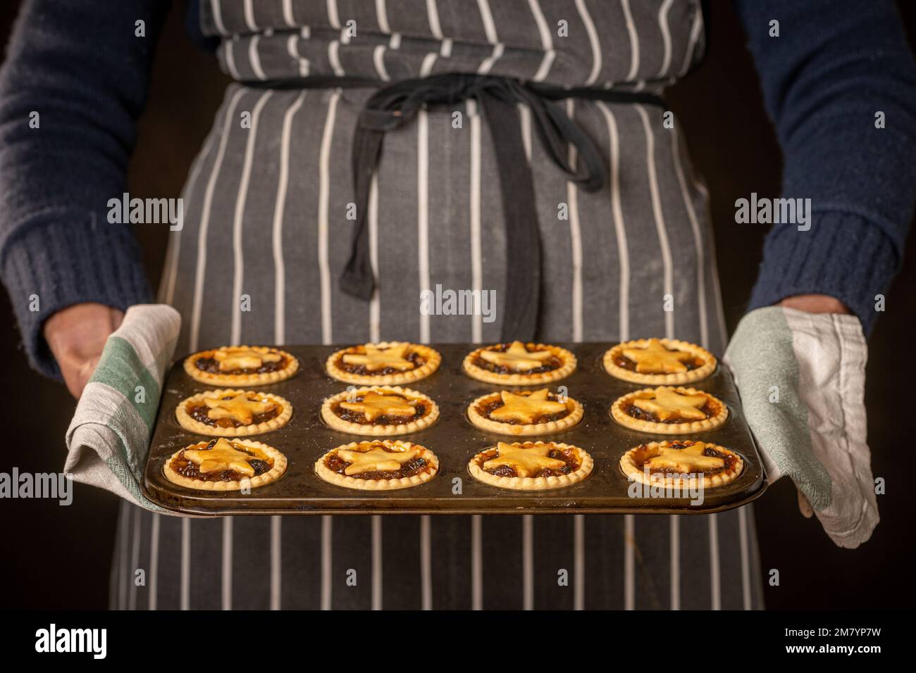 Primo piano delle mani di una persona non identificabile che indossa un tradizionale grembiule a strisce, che tiene un vassoio di torta calda, sormontata da stelle, con un tovagliolo da tè. Foto Stock