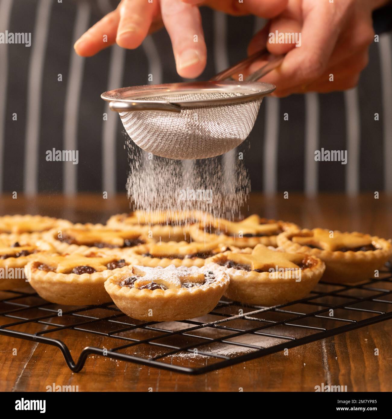 Torte tritate sormontate da stelle di pasta su una griglia metallica che viene dragata in zucchero a velo da una persona non identificabile in un grembiule a strisce nere. Foto Stock