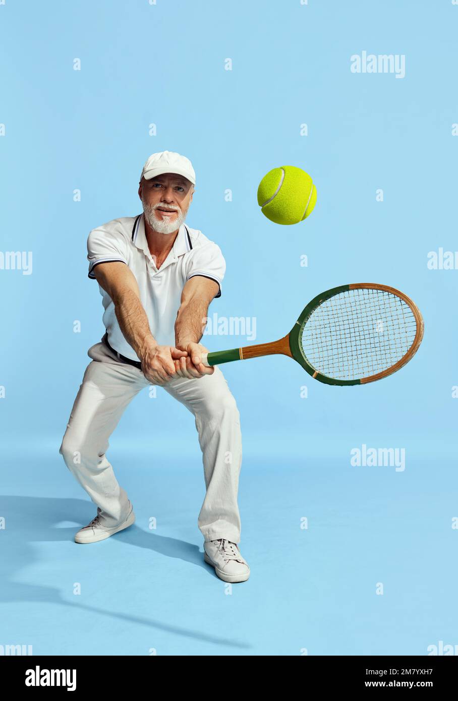 Palla di servizio. Ritratto di bell'uomo anziano in elegante vestito bianco giocando a tennis su sfondo blu. Concetto di attività di svago, hobby Foto Stock