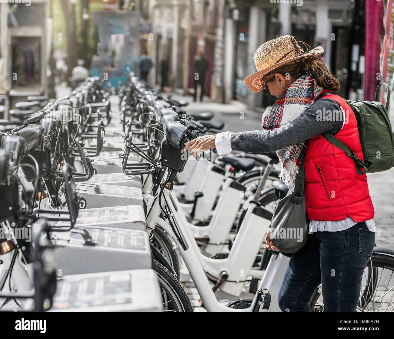 Vista generale di una donna di mezza età che indossa un cappello, scegliendo una bicicletta elettrica per il noleggio. Concetto di trasporto ecologico in città. Foto Stock