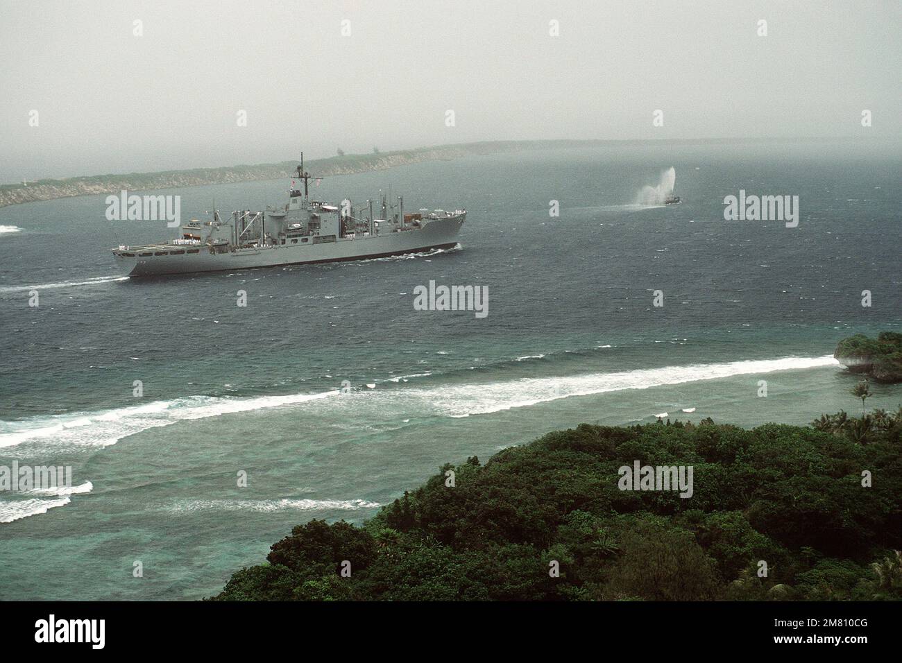 I negozi di combattimento della nave USS NIAGARA FALLS (AFS 3) entrano nel porto di Apra mentre si va verso la stazione navale di Guam. La nave è preceduta da un rimorchiatore che sputa acqua. Il Glas Breakwater artificiale è visibile sullo sfondo. Base: Apra Harbor Stato: Guam (GU) Nazione: Isole Marianne Settentrionali (MNP) Foto Stock