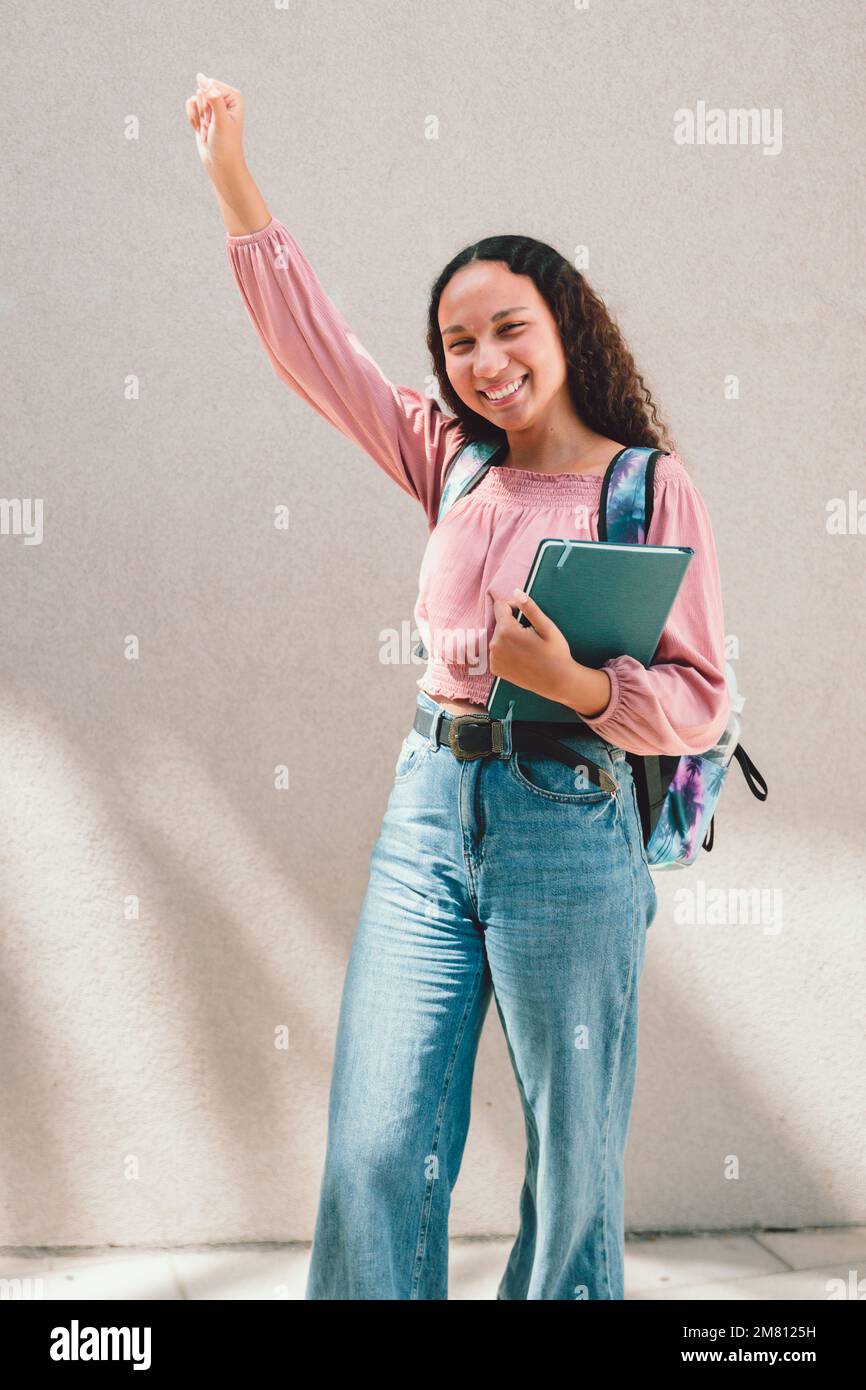 Ragazza studentesca nera entusiasta sollevando il braccio e tenendo i libri contro un muro. Concetto di istruzione Foto Stock