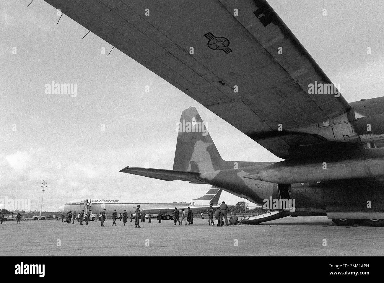 Una polizia di sicurezza dell'aviazione militare statunitense sta a guardia mentre i cittadini cubani vengono trasferiti da un aereo C-130 Hercules a un aereo di linea cubano I1-62M che li riporterà a Cuba. Sono stati catturati a Grenada durante il multiservizio, operazione multinazionale FURIA URGENTE. Soggetto operativo/Serie: URGENT FURY base: Seawell International Airport Paese: Bangladesh (BGD) Foto Stock