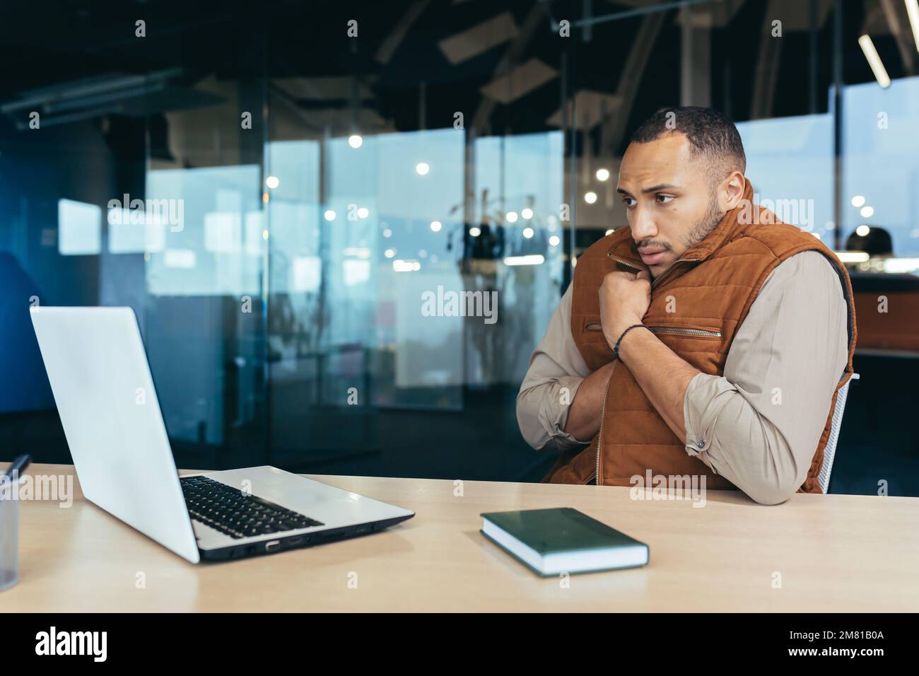 Uomo afroamericano che congela in ufficio, non riscalda sul posto di lavoro, uomo in outerwear seduto alla scrivania con un computer portatile al lavoro. Foto Stock