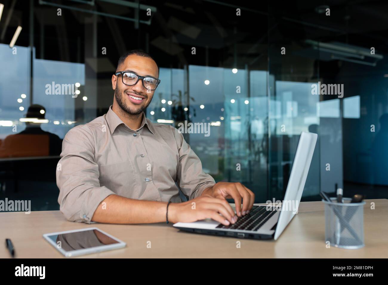 Ritratto di un uomo d'affari ispanico di successo all'interno dell'ufficio, uomo con un computer portatile che lavora a digitare sulla tastiera sorridendo e guardando la fotocamera. Foto Stock