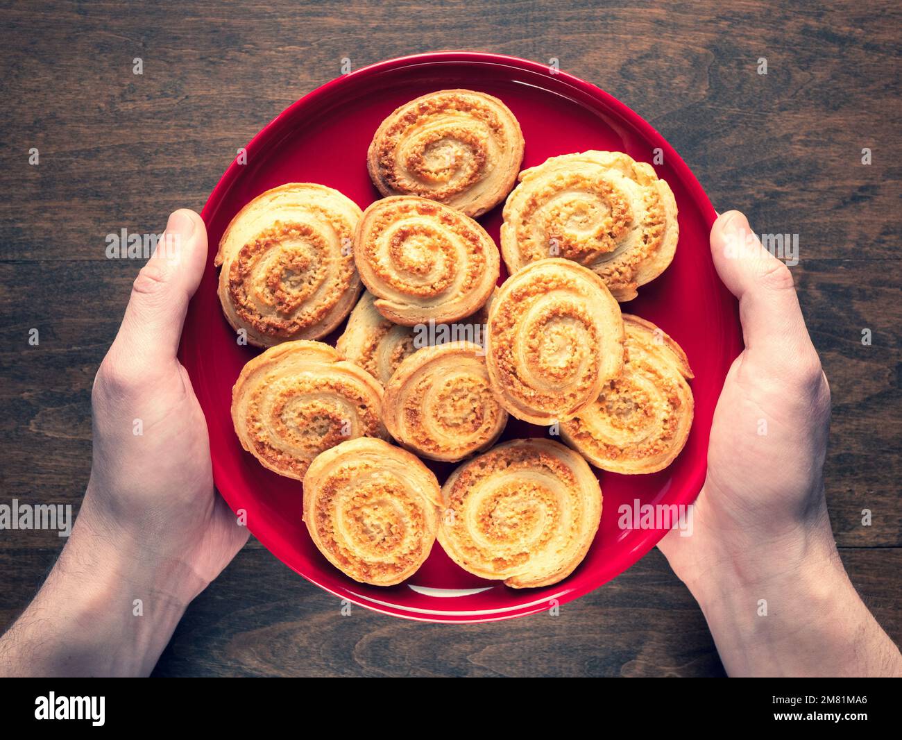 Biscotti flaky deliziosamente croccanti a forma di spirale su un piatto rosso in mani umane, fuoco selettivo. Biscotti fatti in casa con arlette, vista dall'alto Foto Stock