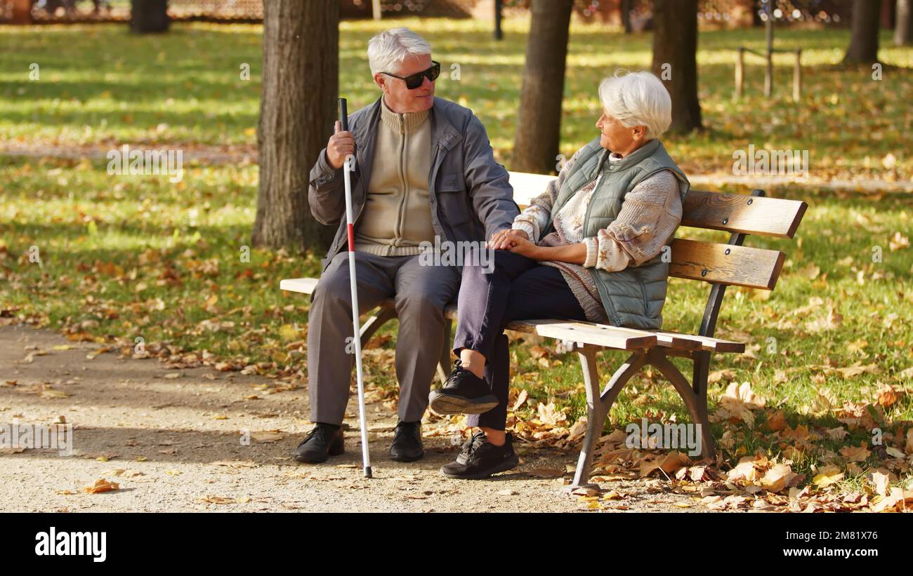 Uomo anziano cieco con occhiali da sole seduto con la moglie su una panchina nel parco. Foto di alta qualità Foto Stock