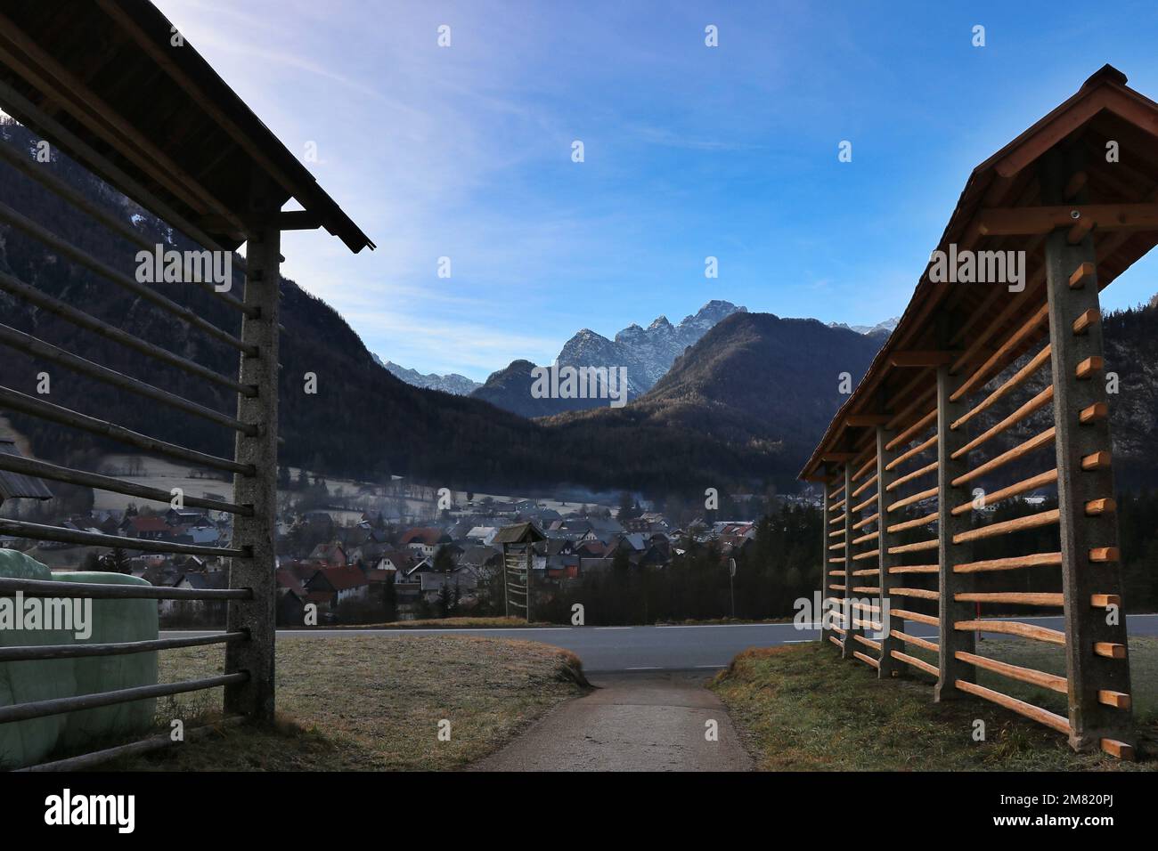 Tradizionale fienagione slovena, o kozolec, sopra il villaggio di Mojstrana immerso nella valle sotto le Alpi Giulie e il Monte Triglav Foto Stock
