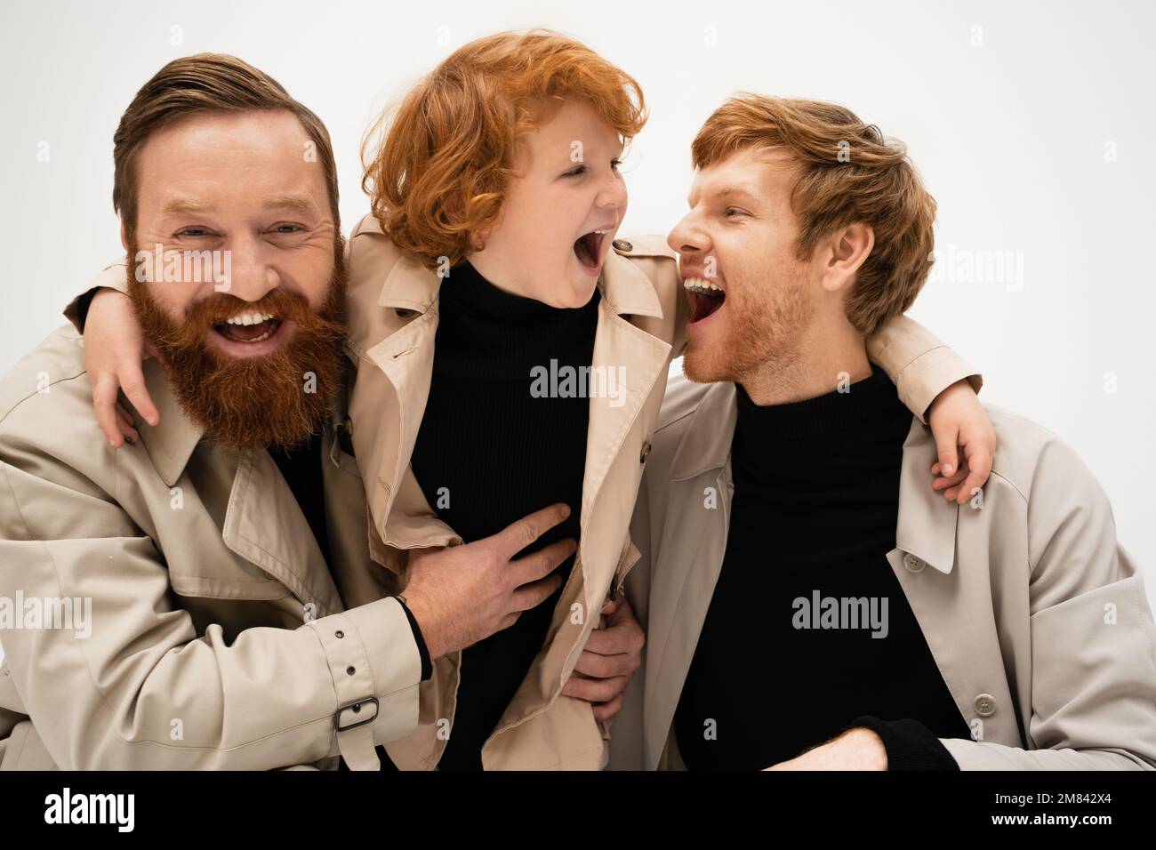 gioiosi uomini bearded e ragazzo con capelli rossi in trench cappotti ridendo isolato su grigio, immagine stock Foto Stock