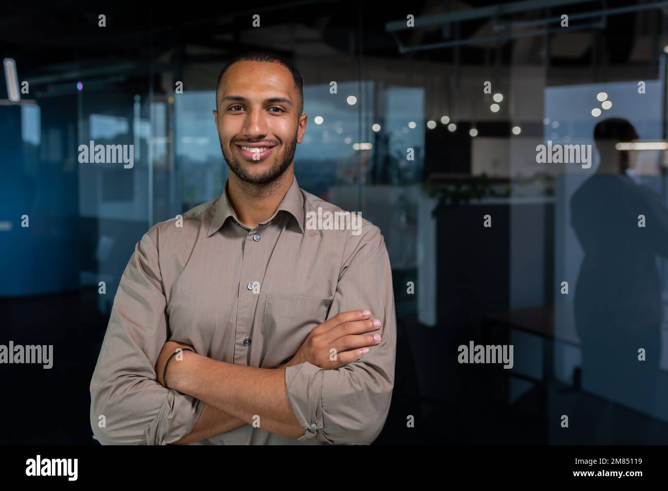 Ritratto di uomo d'affari ispanico riuscito, uomo riuscito in camicia che guarda la macchina fotografica con le braccia incrociate, operaio all'interno dell'ufficio vicino alla finestra. Foto Stock