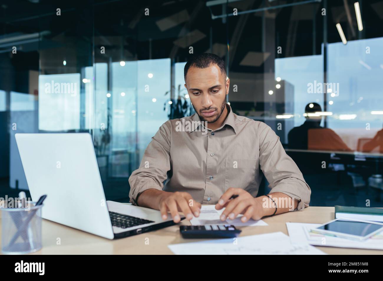 Serio uomo d'affari ispanico che lavora con documenti e fatture all'interno dell'ufficio, uomo dietro lavoro di carta, usando il laptop al lavoro seduto alla scrivania. Foto Stock