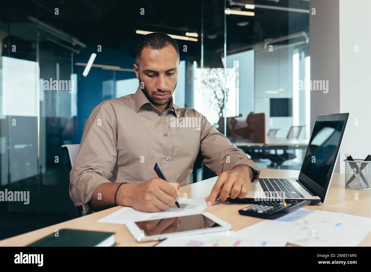 Serio uomo d'affari ispanico che lavora con documenti e fatture all'interno dell'ufficio, uomo dietro lavoro di carta, usando il laptop al lavoro seduto alla scrivania. Foto Stock