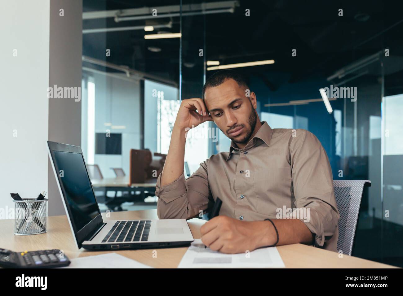 Serio uomo d'affari ispanico che lavora con documenti e fatture all'interno dell'ufficio, uomo dietro lavoro di carta, usando il laptop al lavoro seduto alla scrivania. Foto Stock