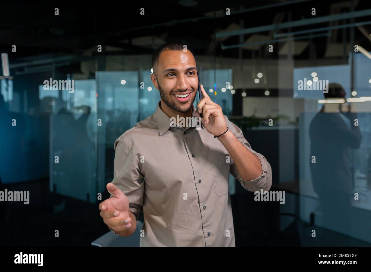 Uomo d'affari allegro e sorridente in una camicia che parla al telefono all'interno dell'ufficio, uomo ispanico in piedi vicino alla finestra di sera. Foto Stock