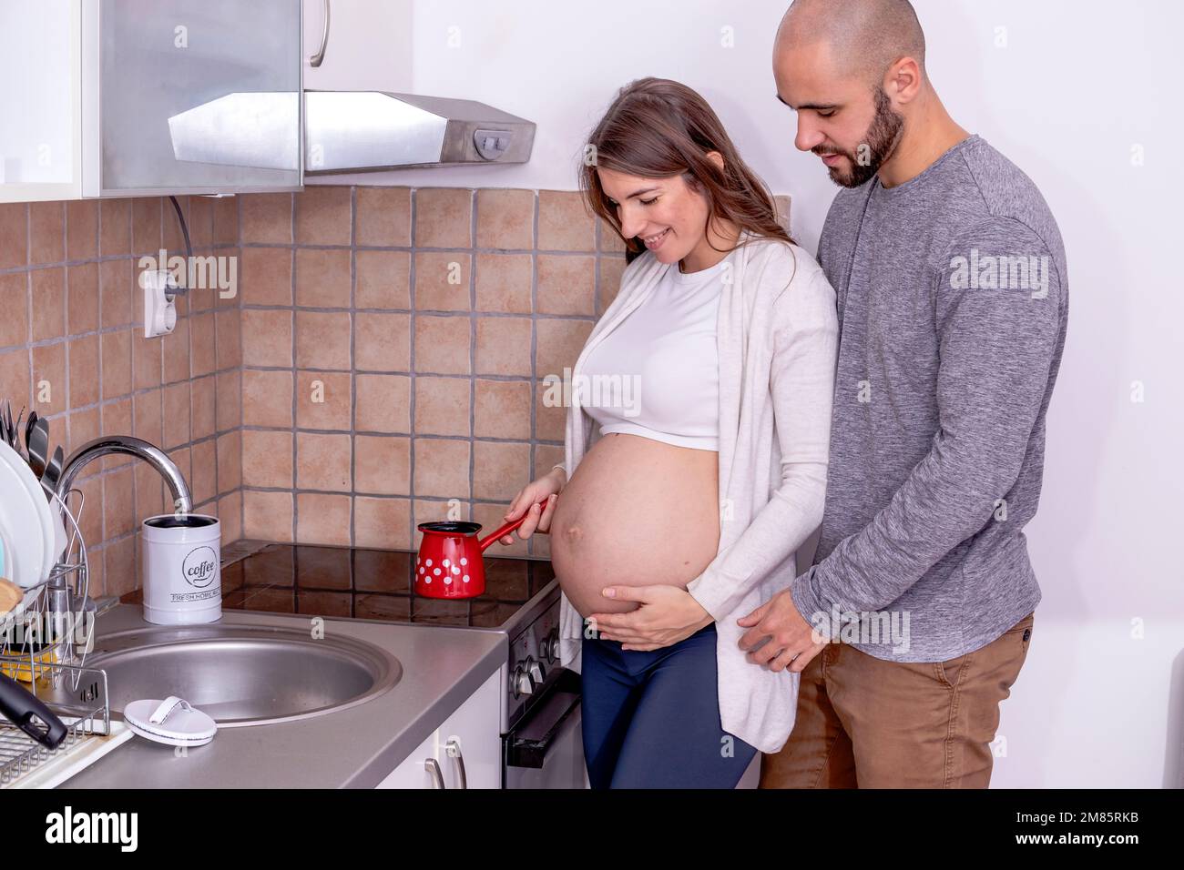 Una giovane donna incinta che fa il caffè in cucina, con il marito che la sostiene. Foto Stock