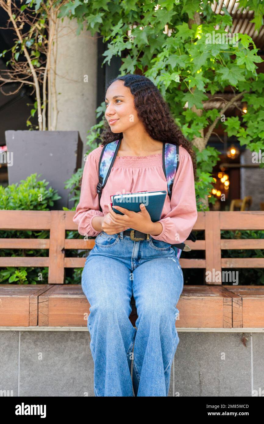 Donna studentesca nera sorridente seduta e in attesa di un compagno di classe fuori dal centro commerciale. Concetto di istruzione Foto Stock