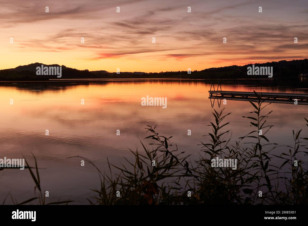 Wundervolle Abendstimmung am Hopfensee mit Steg und orangeroter Wollenspiegelung im See Foto Stock