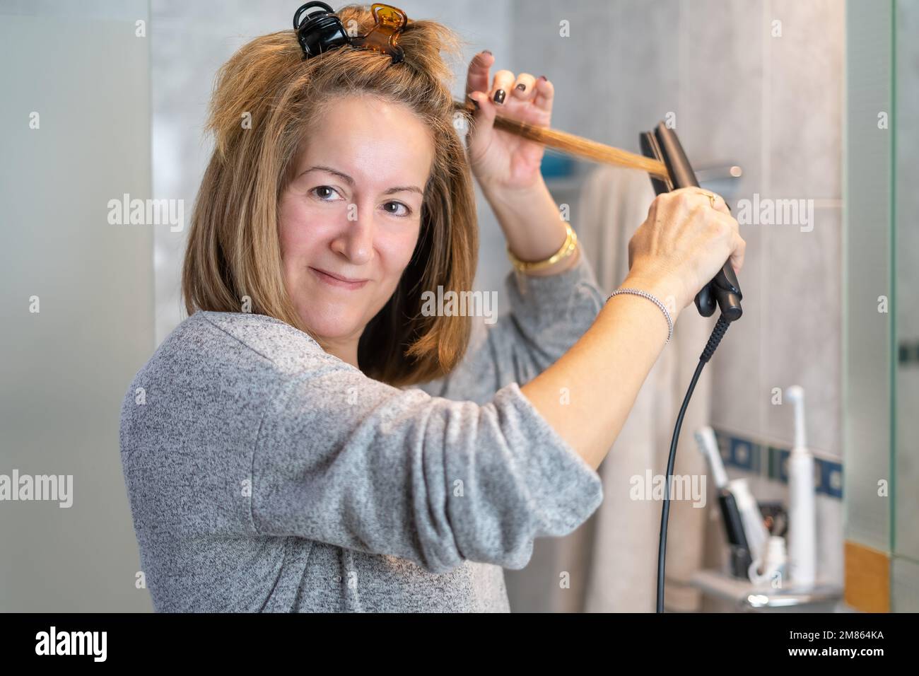 Donna bianca matura fissando i capelli davanti allo specchio con un apparecchio per lisciare i capelli. Foto Stock