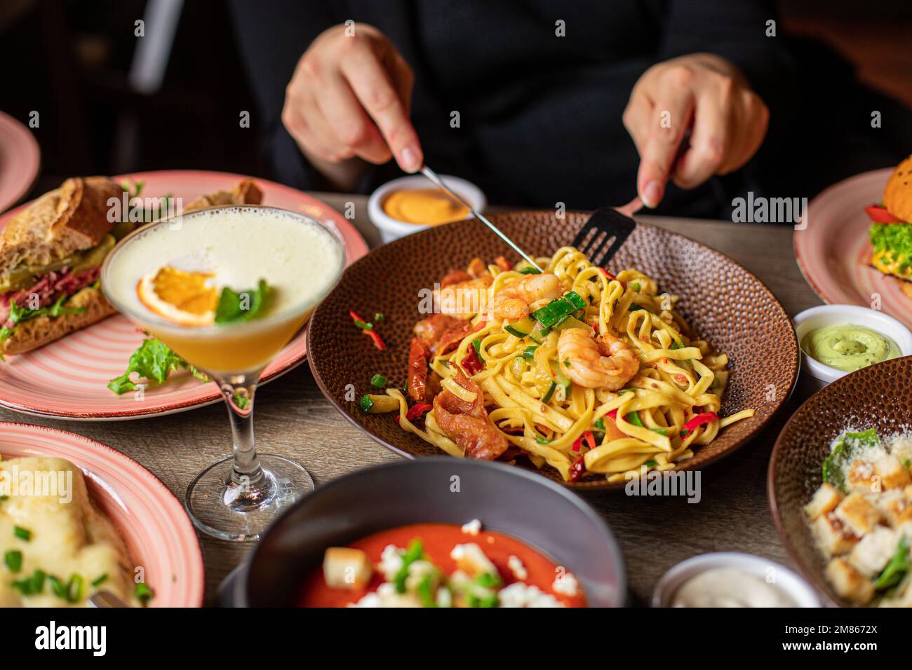 Umano con forchetta e coltello mangiare pasta con gamberetti dal piatto su tavolo pieno di cibo delizioso. Piatti caldi e bevande alcoliche durante la cena festiva Foto Stock