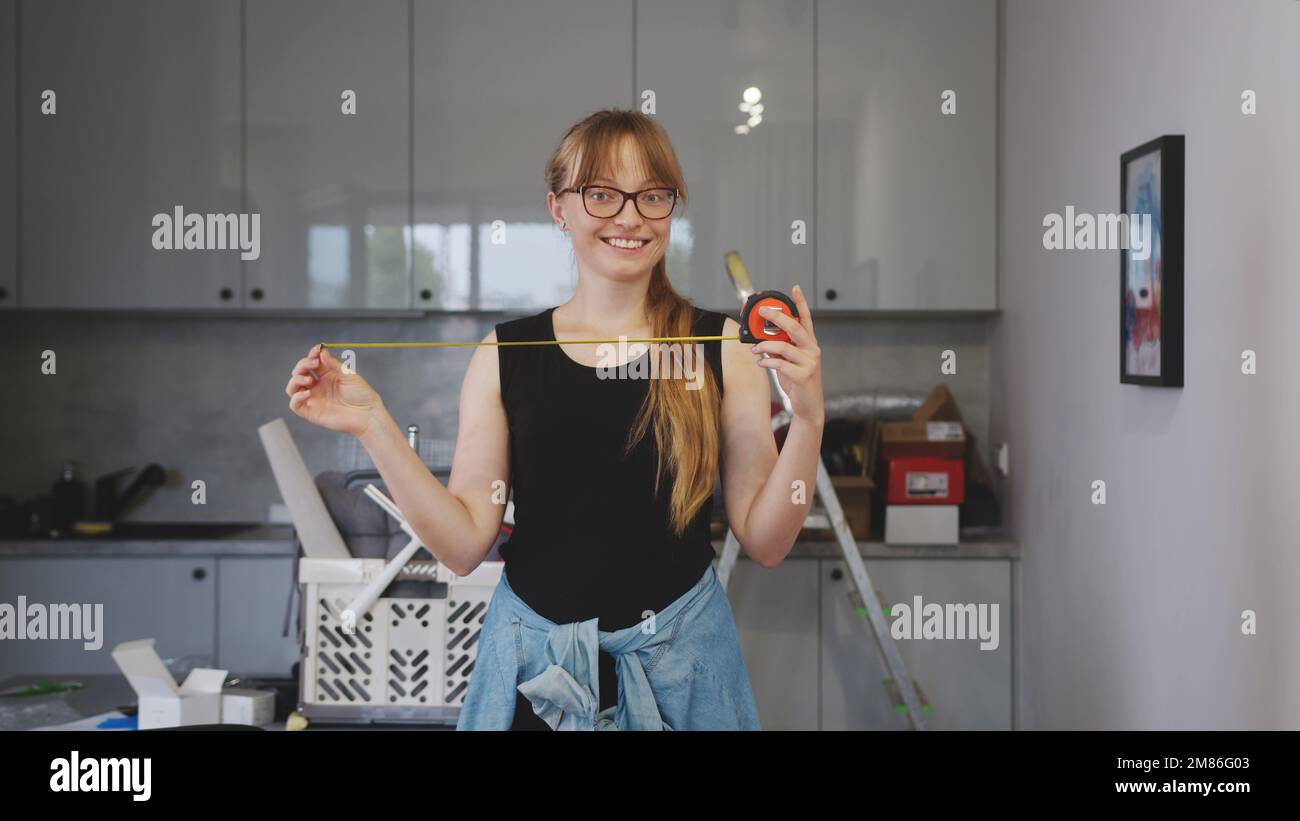 donna felice e soddisfatta che tiene un nastro di misurazione aperto in cucina, attrezzi dietro di lei. Foto di alta qualità Foto Stock