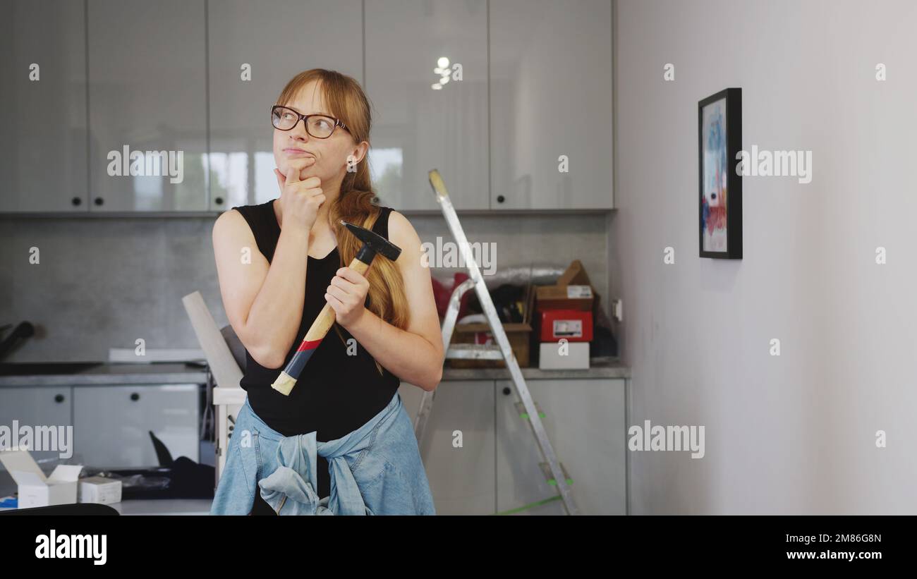 donna dai capelli biondi con un martello in mano pensando alla ristrutturazione in cucina, utensili in background. Foto di alta qualità Foto Stock