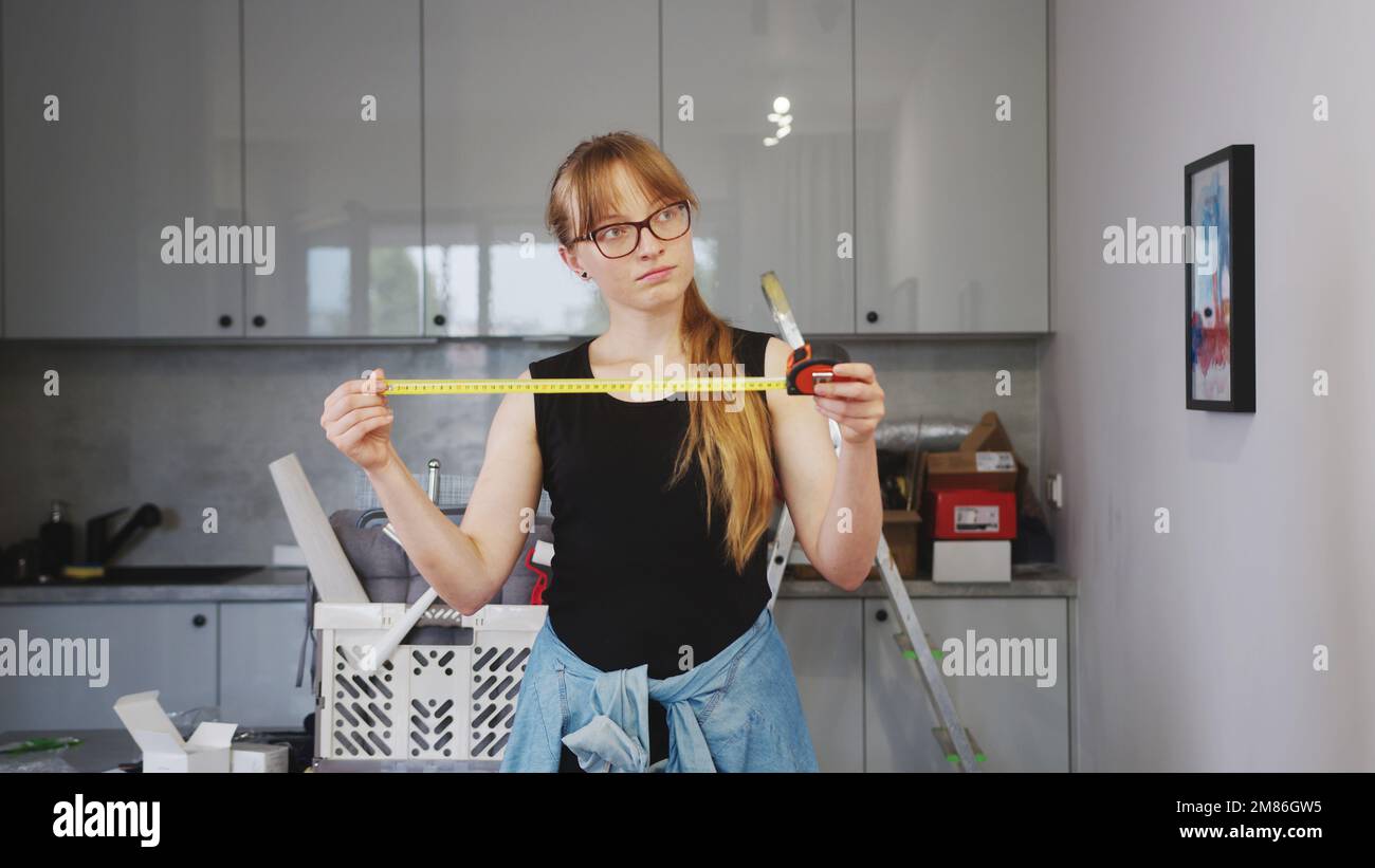 tempo di rinnovamento, giovane donna in camicia nera senza maniche con lunghi capelli biondi e bicchieri che tengono un metro a nastro in cucina. Foto di alta qualità Foto Stock