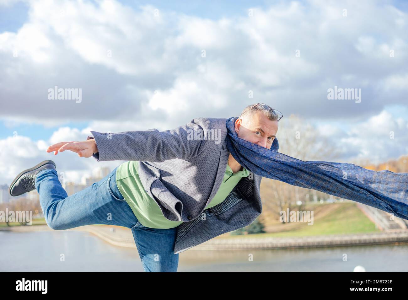 Divertente uomo grizzled maturo in abiti caldi in piedi su una gamba con la gamba sollevata e le braccia sul bordo del parco in una giornata ventosa Foto Stock