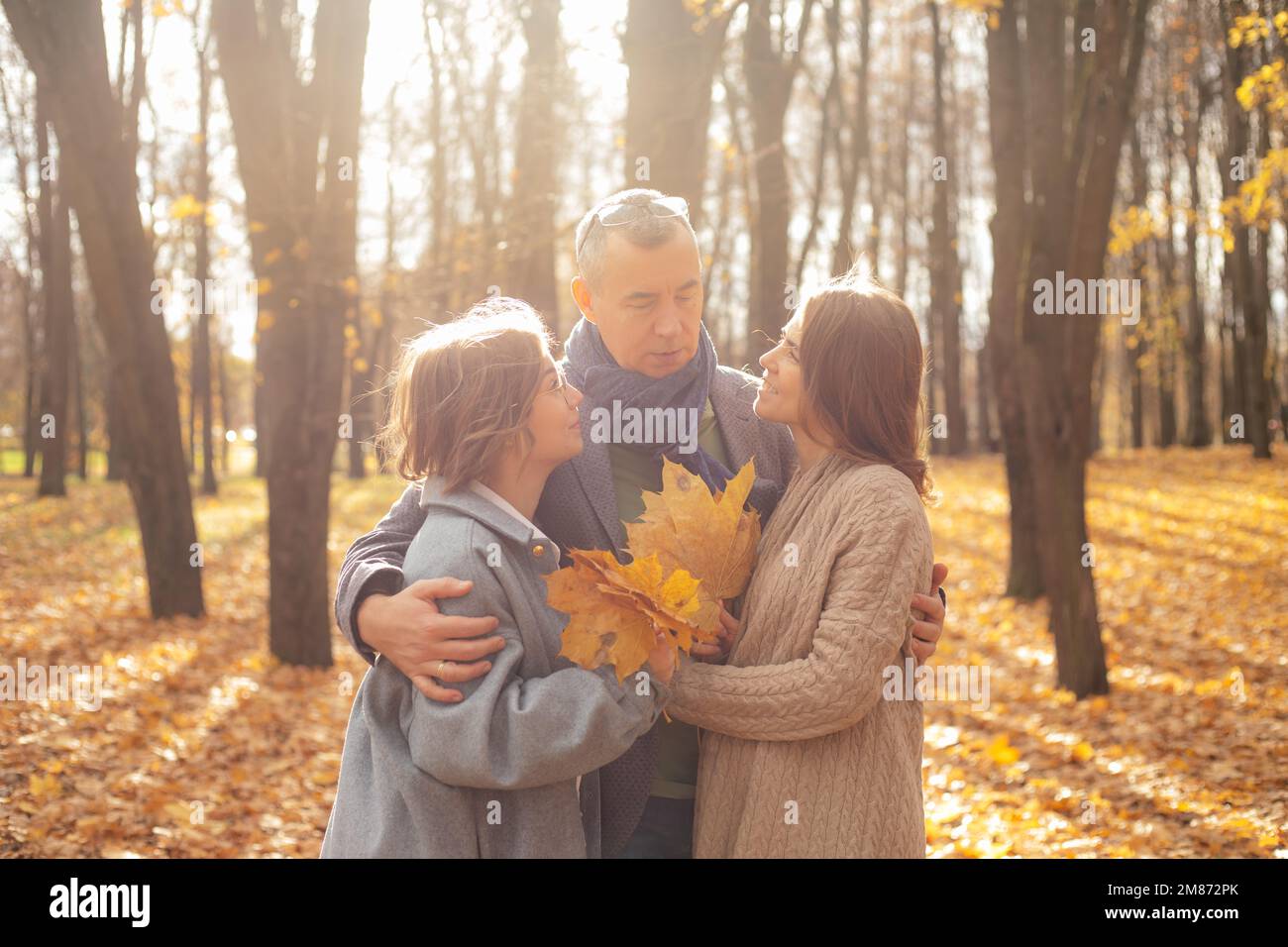 Deliziosa famiglia di madre, padre, figlia divertirsi insieme, abbracciare, raccogliere le foglie e rilassarsi nei boschi dorati Foto Stock