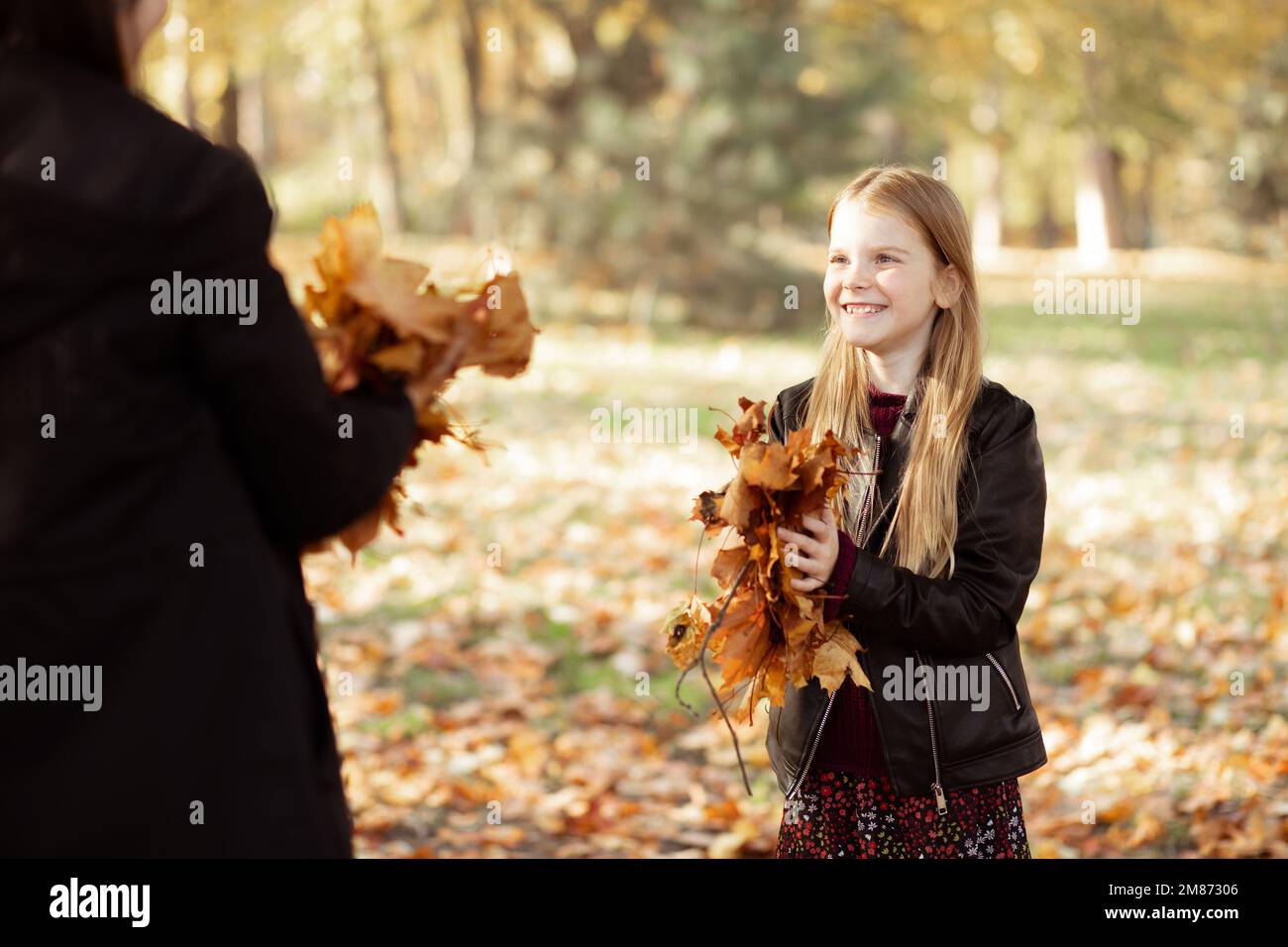 Primo piano sorridente ragazza bionda felice, spensierata in giacca nera giocare e raccogliere le foglie in autunno foresta dorata Foto Stock
