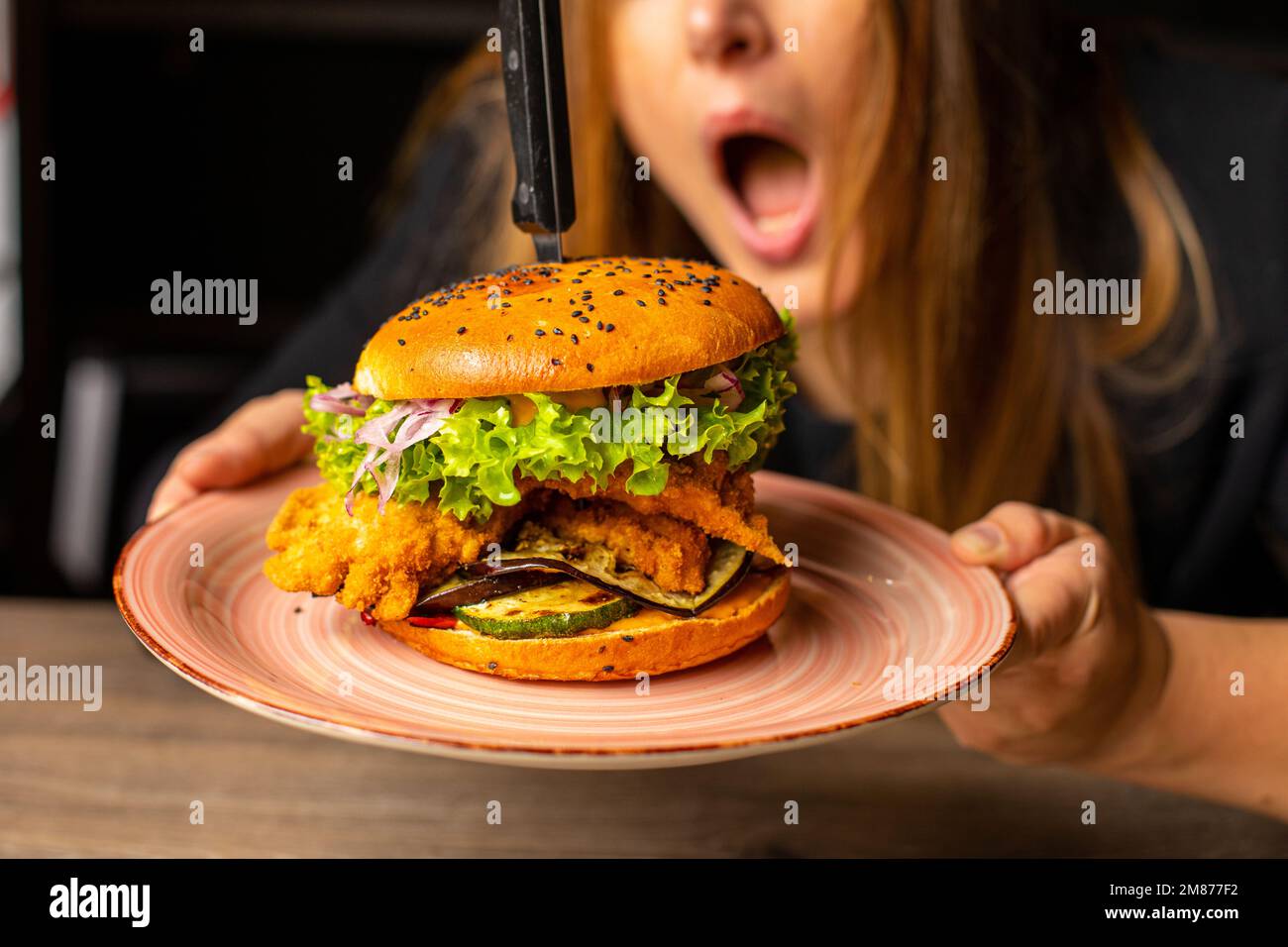 Foto ritagliata di una donna scioccata con bocca aperta che tiene gustoso hamburger con lattuga, cipolla rossa, pomodoro, pollo fritto. Foto Stock