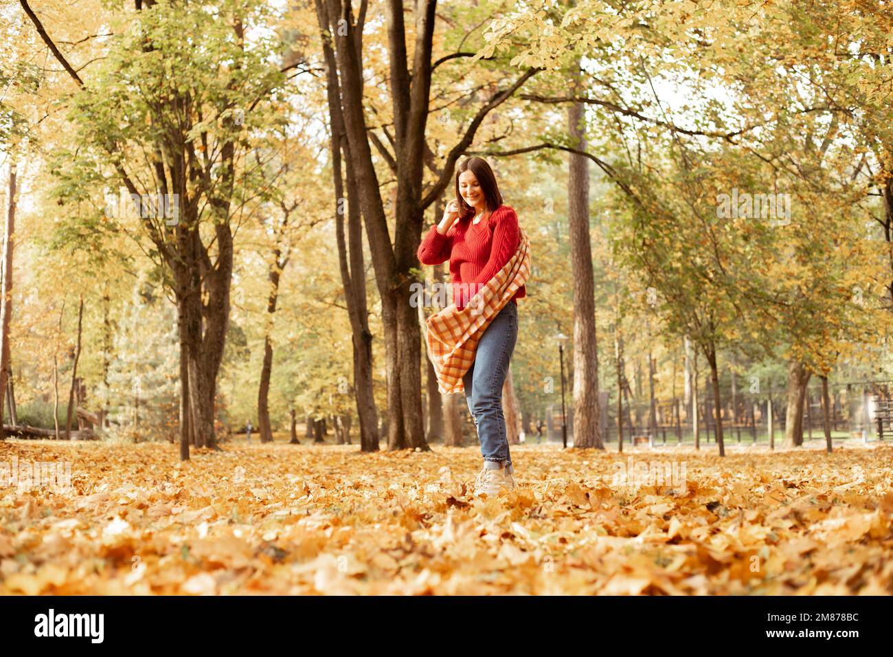 Donna sorridente, elegante dai capelli scuri in una calda sciarpa a pipa e un comodo maglione rosso che posa e cammina nella foresta delle foglie dorate Foto Stock