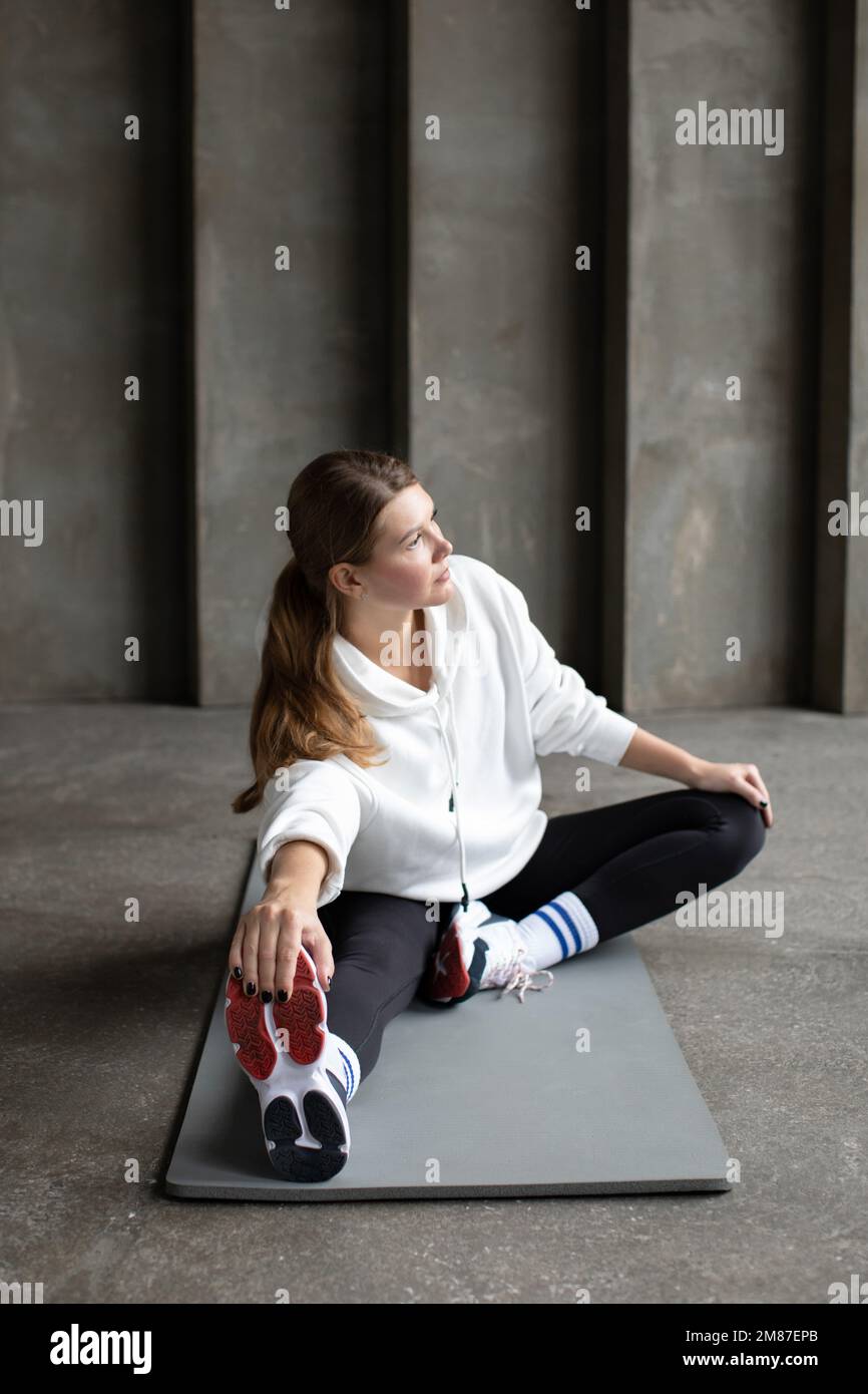 Un atleta con leggings, felpa con cappuccio e sneaker allunga i muscoli delle gambe prima di allenarsi Foto Stock