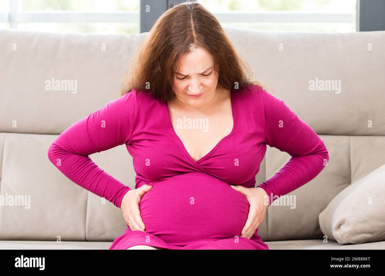 Ritratto di una donna incinta che soffre mal di pancia seduto su un divano nel soggiorno in una casa interna con una luce calda sullo sfondo Foto Stock