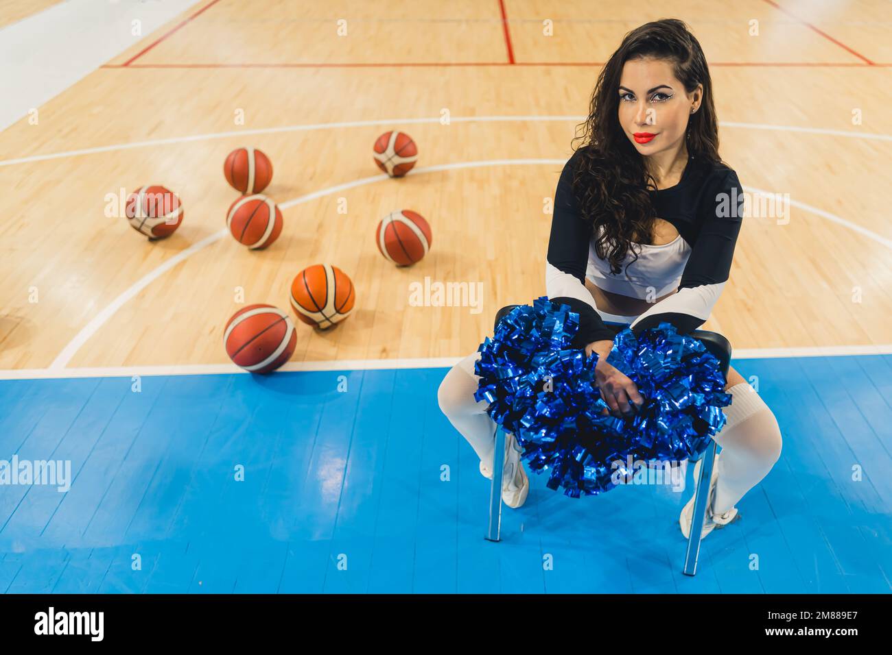 Un cheerleader attraente siede con fiducia, tenendo i suoi pom-pom. È vestita con una camicia nera e calzini alti. Un senso di potenza ed energia. Dietro di lei si può vedere un mucchio di palline da basket. Foto Stock