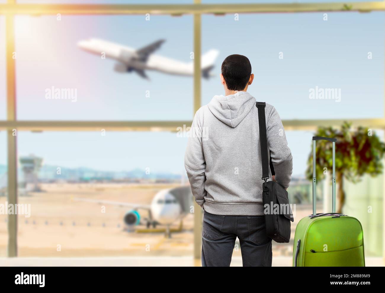 Il turista maschile calmo è in piedi in aeroporto e guardando il volo aereo attraverso la finestra. Ha in mano biglietti e valigia. Tramonto Foto Stock