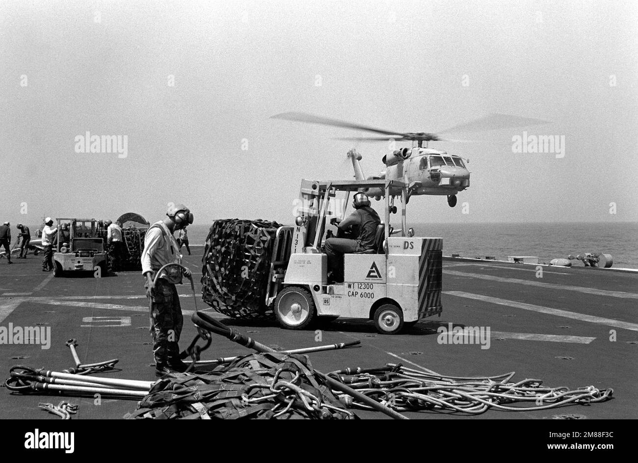 Un elicottero SH-60B Sea Hawk atterra sullo sfondo mentre gli equipanti rimuovono pallet di rifornimenti dal ponte di volo della nave d'assalto anfibia USS GUADALCANAL (LPH-7). Paese: Golfo di Oman Foto Stock